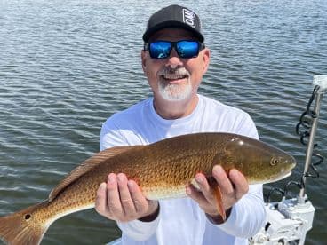 An angler proudly displaying a Redfish while fishing on a sunny day, showcasing the vibrant colors of the catch against the water backdrop.