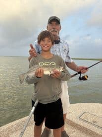 A young angler proudly displaying a Speckled Trout while fishing with an adult on a boat in a coastal environment.