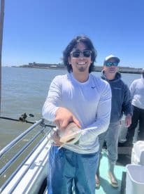 A young angler proudly displaying a caught fish while fishing on a boat, with a sunny backdrop and fellow anglers in the background.