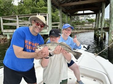 A family proudly displaying a large Speckled Trout on a boat, showcasing a successful fishing trip in a scenic waterway.