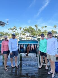 A group of four anglers proudly displaying their catch of the day, featuring a variety of fish, including a large grouper, on a fish rack at the marina.