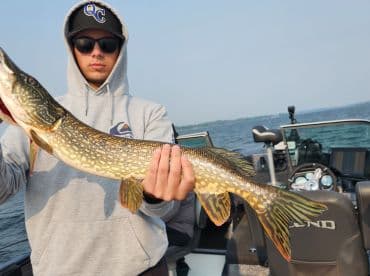 Angler displaying a large Northern Pike while fishing from a Lund boat on a calm lake.