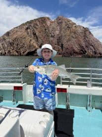A young angler proudly displaying a striped bass while fishing near rocky cliffs on a sunny day.