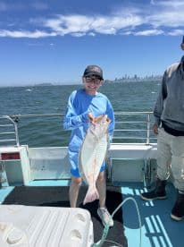 A young angler proudly displaying a large Halibut while fishing on a boat in a scenic bay, with the city skyline in the background.
