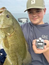 Young angler proudly holding a large Bass while fishing on a boat, showcasing a successful catch in a serene water environment.