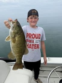 A young angler proudly displaying a large Smallmouth Bass while fishing on a calm lake.