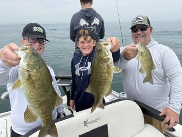 A family fishing trip showcasing three anglers proudly holding their catch of Smallmouth Bass while on a boat.