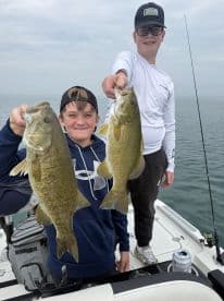 Two young anglers proudly displaying their catch of large Smallmouth Bass while fishing on a boat.