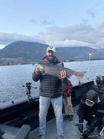 An enthusiastic angler proudly displaying a large lake trout while fishing on a scenic lake surrounded by mountains.