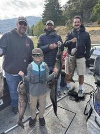 A group of anglers, including a young boy, proudly displaying their catch of salmon on a fishing boat in a scenic lake setting.