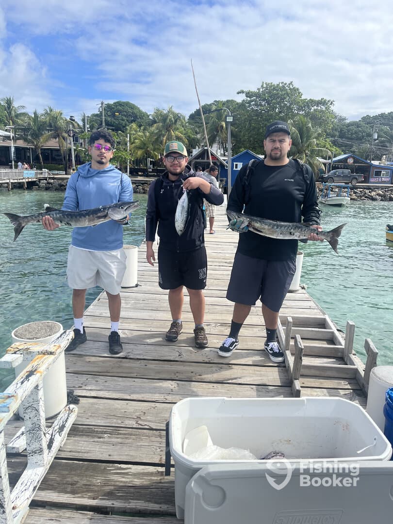 Three anglers proudly displaying their catch, including a large Barracuda and a smaller fish, on a dock in a tropical fishing environment.