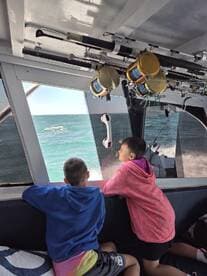 Two young anglers enjoying the view from inside the boat, with fishing rods visible above them, as they head out to sea.