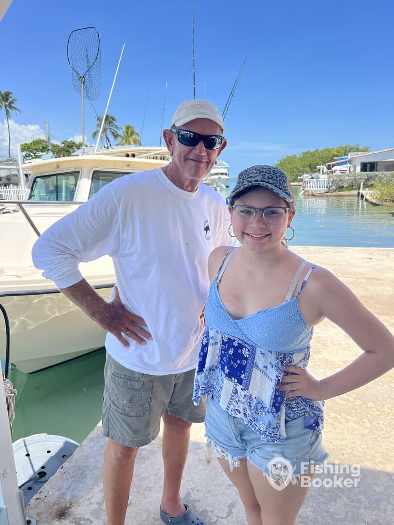 A man and a young girl pose together at the dock, with a fishing boat in the background, suggesting a day of fishing ahead.