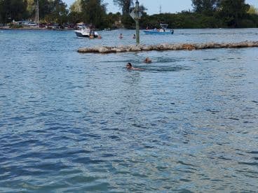 A scenic view of swimmers enjoying the clear waters near a rocky shoreline, with boats in the background, capturing a vibrant day at the beach.