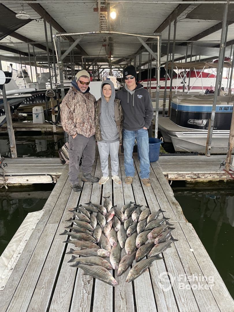 A group of three anglers proudly posing with a large catch of striped bass arranged in a heart shape on the dock, showcasing their successful fishing trip.